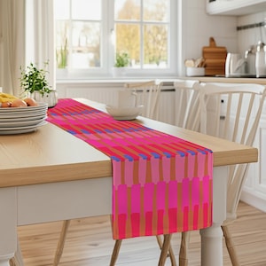 Könnte beinhalten: Ein Tischläufer mit geometrischem Muster in Pink, Orange und Blau. Der Läufer liegt auf einem Holztisch mit zwei weißen Stühlen.