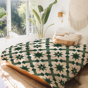 May include: A bed with a cream-colored comforter featuring a geometric pattern of dark green stars and diamond shapes. The bed is made of wood and is in a bright, airy room with a large window and a houseplant.