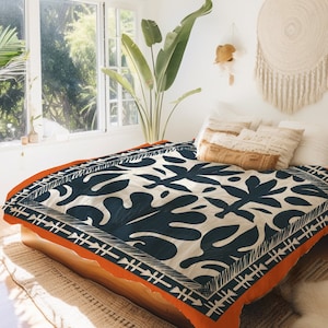 Op de afbeelding: Een bed met een wit en blauw bedrukt dekbedovertrek met een oranje rand. Het dekbedovertrek heeft een herhalend patroon van gestileerde bladeren. Het bed is opgemaakt met kussens en een deken.