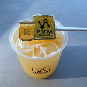 Puede incluir: Un vaso de plástico transparente lleno de una bebida de color amarillo con cubitos de hielo. Un pin de metal dorado con el texto "PYM Tasting Lab" y un patrón de panal de abeja está unido a una pajita blanca.