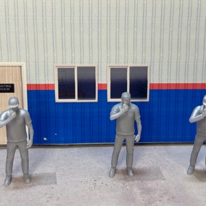 May include: Three gray plastic figures of people wearing hats and standing in front of a building with a blue and white striped wall. The figures are all looking down and have their hands on their chins.