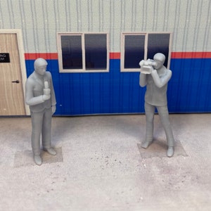 May include: Two gray plastic figurines of reporters, one holding a microphone and the other holding a video camera. The figures are standing on a gray surface in front of a building with a red and blue striped wall.