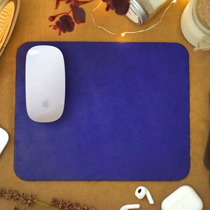 Peut inclure: Un tapis de souris en cuir violet avec une souris sans fil blanche sur le dessus. Le tapis de souris est sur une surface brune avec d'autres objets autour.