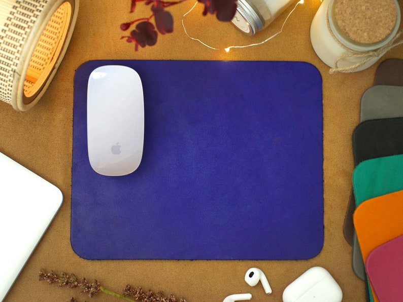 Peut inclure: Un tapis de souris en cuir violet avec une souris sans fil blanche sur le dessus. Le tapis de souris est sur une surface brune avec d'autres objets autour.