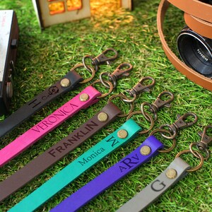 May include: Six colourful leather camera straps with metal clasps and engraved names: Veronica, Franklin, Monica, ARV, and G. The straps are arranged on a green grassy surface.
