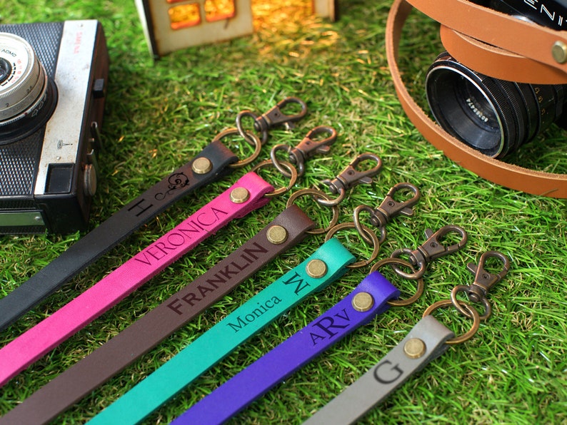 May include: Six colourful leather camera straps with metal clasps and engraved names: Veronica, Franklin, Monica, ARV, and G. The straps are arranged on a green grassy surface.