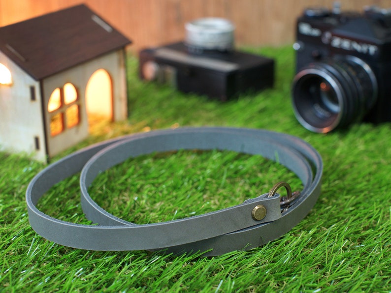 May include: A grey leather camera strap with a brass buckle and a metal ring. The strap is lying on a green grassy surface.