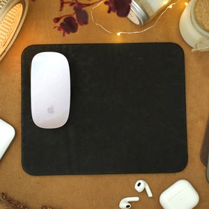 Peut inclure: Un tapis de souris noir avec une souris sans fil blanche sur le dessus. Le tapis de souris est sur une surface brune avec un ordinateur portable blanc, une petite lampe en bois et un bocal blanc avec un couvercle en li&egrave;ge.