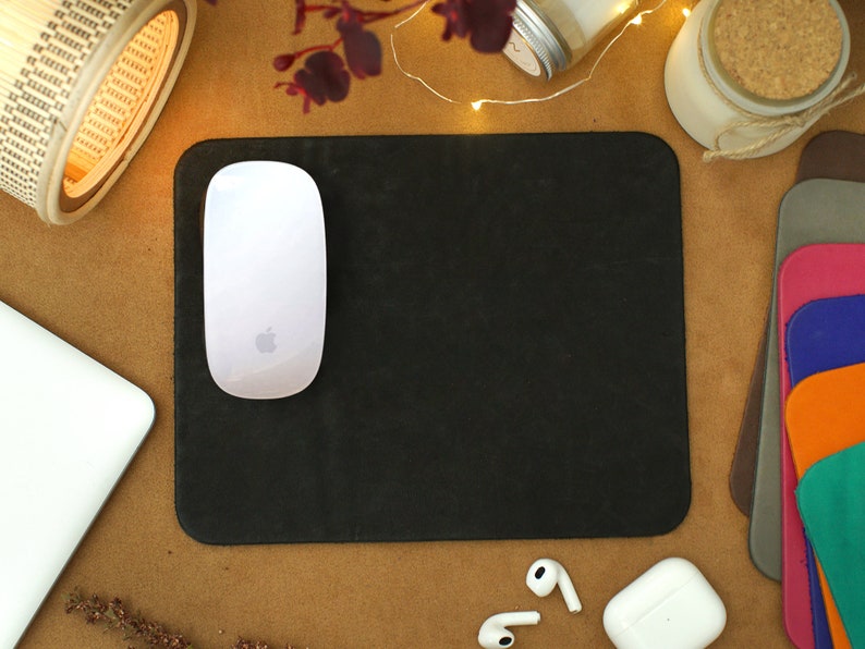 Peut inclure: Un tapis de souris noir avec une souris sans fil blanche sur le dessus. Le tapis de souris est sur une surface brune avec un ordinateur portable blanc, une petite lampe en bois et un bocal blanc avec un couvercle en li&egrave;ge.