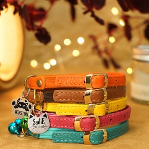 May include: A stack of five colourful leather cat collars with gold buckles. The collars are in shades of orange, brown, yellow, pink, and teal. The top collar has a silver cat paw charm with the name "Winston" engraved on it. The second collar has a silver cat head charm with the name "Sadie" engraved on it. The collars are accented with silver bells.