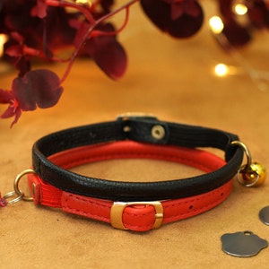 May include: A red and black leather cat collar with a gold buckle and two bells. The collar is lying on a brown surface.