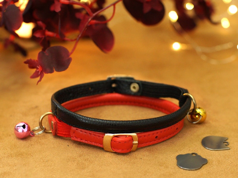 May include: A red and black leather cat collar with a gold buckle and two bells. The collar is lying on a brown surface.