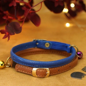 May include: A blue and brown leather cat collar with a gold buckle and two bells. The collar is on a brown surface.