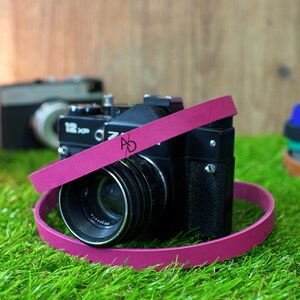 May include: A pink camera strap with the letters "AD" printed on it. The strap is wrapped around a black camera with a lens.