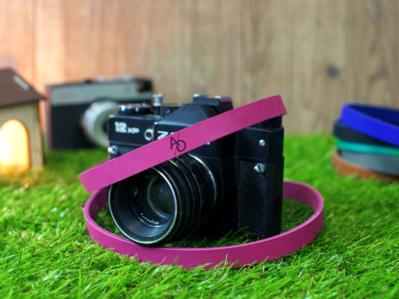 May include: A pink camera strap with the letters "AD" printed on it. The strap is wrapped around a black camera with a lens.