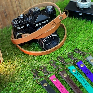 May include: A brown leather camera strap with the name "MARCUS" engraved on it. The strap is wrapped around a black camera with a lens. Five additional leather camera straps with different names engraved on them are shown in the background.