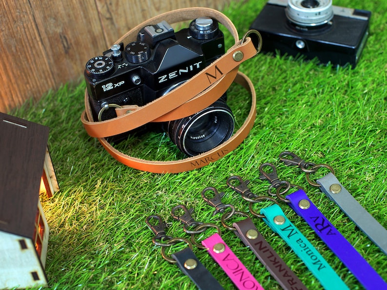 May include: A brown leather camera strap with the name "MARCUS" engraved on it. The strap is wrapped around a black camera with a lens. Five additional leather camera straps with different names engraved on them are shown in the background.