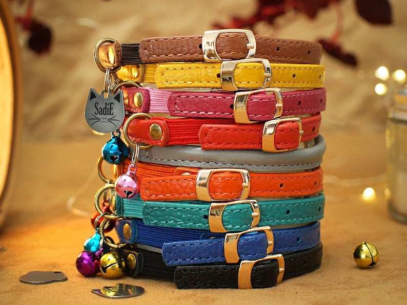 May include: A stack of colourful cat collars with a bell and a tag that says "Sadie" on it. The collars are made of leather and come in a variety of colours, including brown, yellow, pink, red, grey, orange, teal, blue, and black.