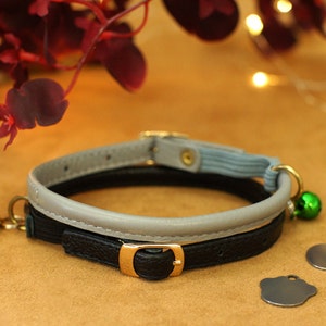 May include: A black and grey cat collar with a grey leather band and a black leather band with a gold buckle. The collar has a green bell and two silver tags.