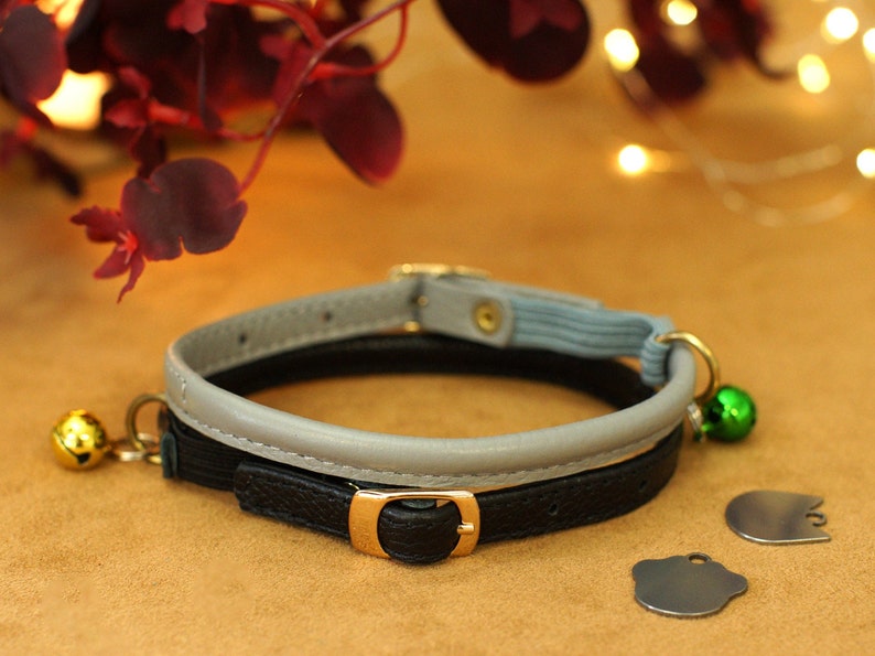 May include: A black and grey cat collar with a grey leather band and a black leather band with a gold buckle. The collar has a green bell and two silver tags.
