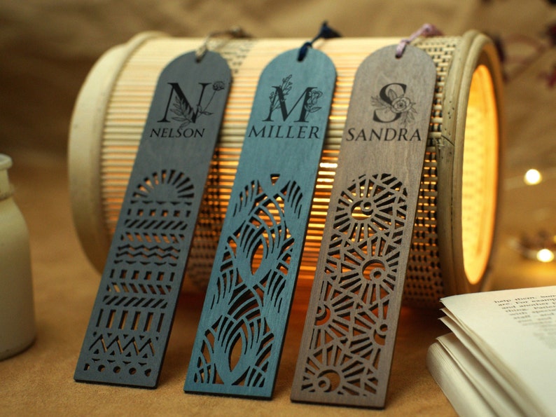 May include: Three wooden bookmarks with laser-cut designs. The bookmarks are dark blue, light blue, and brown. Each bookmark has a letter, a name, and a floral design. The bookmarks are on a bamboo surface.