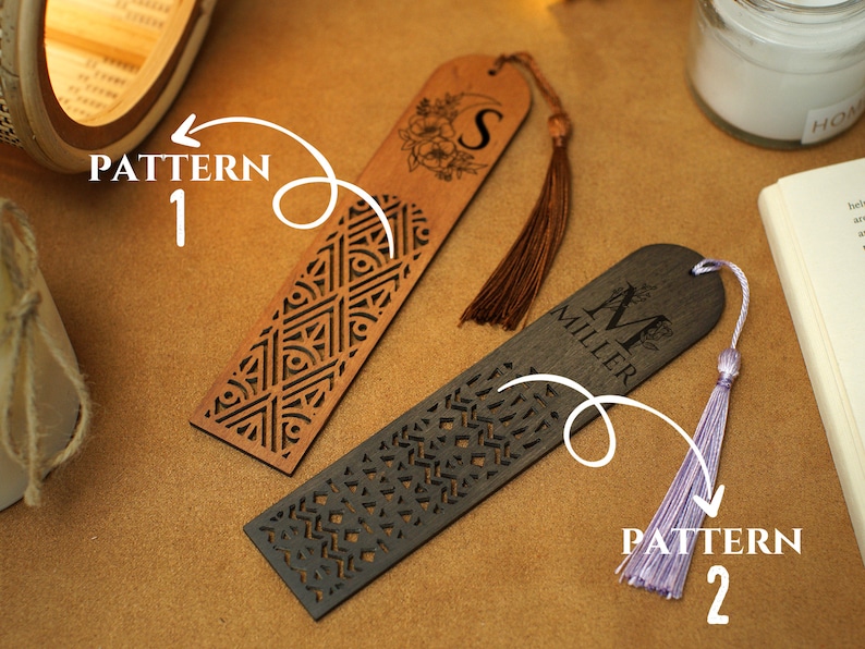 May include: Two wooden bookmarks with different patterns and tassels. The bookmark on the left has a geometric pattern and a brown tassel. The bookmark on the right has a floral pattern and a purple tassel. The bookmark on the right has the name "Miller" and the initial "M" engraved on it.