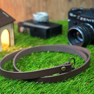 May include: A brown leather camera strap with a metal ring and a small metal button. The strap is lying on a green grassy surface.