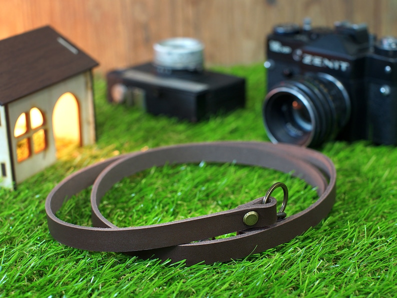 May include: A brown leather camera strap with a metal ring and a small metal button. The strap is lying on a green grassy surface.