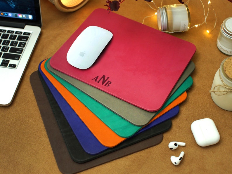 Peut inclure: Une pile de huit tapis de souris en cuir personnalis&eacute;s dans diff&eacute;rentes couleurs, dont le marron, le bleu marine, l'orange, le vert, le gris et le rouge. Le tapis de souris du dessus porte les initiales "ANB" en relief blanc.