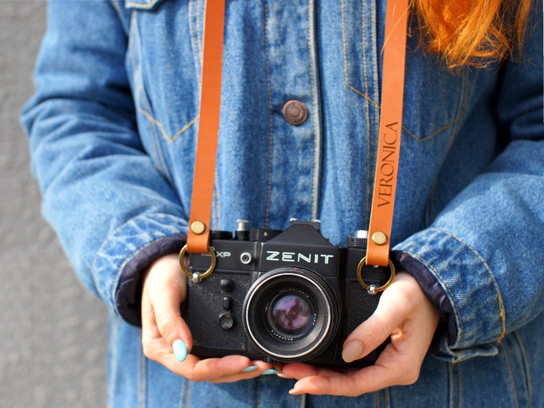 May include: A person holding a black Zenit camera with a brown leather camera strap that says "VERONICA" on it.