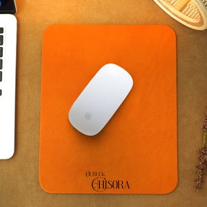 Peut inclure: Un tapis de bureau en cuir orange avec une souris sans fil blanche et le texte "Derek Chisora" en noir.