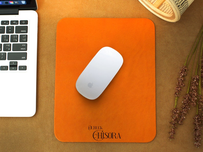 Peut inclure: Un tapis de bureau en cuir orange avec une souris sans fil blanche et le texte "Derek Chisora" en noir.