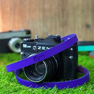 May include: A purple camera strap with the word "Gloria" printed on it. The strap is wrapped around a black camera lens.
