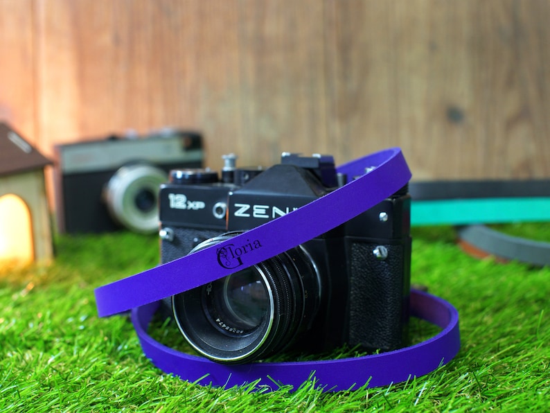 May include: A purple camera strap with the word "Gloria" printed on it. The strap is wrapped around a black camera lens.