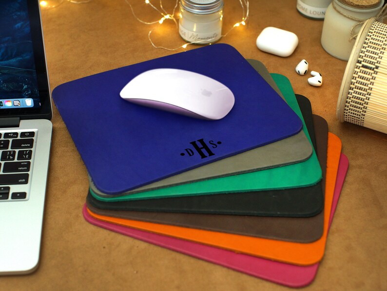 Peut inclure: Une pile de tapis de souris en cuir de diff&eacute;rentes couleurs avec une souris sans fil blanche sur le dessus du tapis de souris bleu. Le tapis de souris du dessus porte les initiales "D.H.S." en lettres noires.