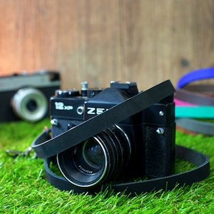 May include: A black vintage camera with a leather strap, sitting on green grass. The camera has a lens cap and the text "122 KCP" and "OZE" on the body.