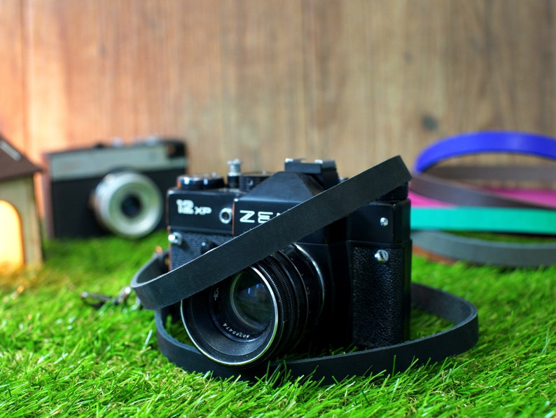 May include: A black vintage camera with a leather strap, sitting on green grass. The camera has a lens cap and the text "122 KCP" and "OZE" on the body.