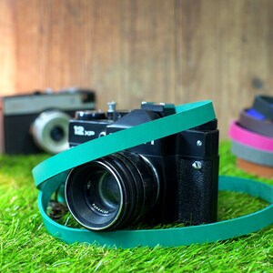 May include: A vintage black camera with a teal camera strap. The camera has a large lens and is sitting on green grass.
