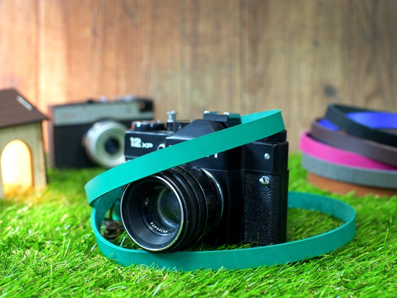 May include: A vintage black camera with a teal camera strap. The camera has a large lens and is sitting on green grass.