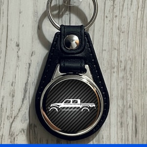 May include: Black leather keychain with a silver round charm featuring a white silhouette of a pickup truck on a carbon fiber background.