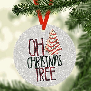 May include: Round Christmas ornament with a glittery silver background. The ornament features the text "OH CHRISTMAS TREE" in a mix of plaid and green lettering. A decorated Christmas tree cookie is on the right side, and a red ribbon is attached for hanging.