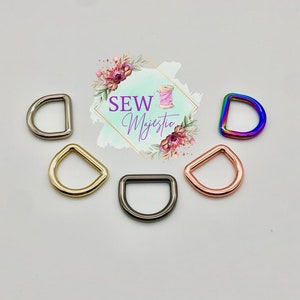 Puede incluir: Cinco anillas en D de diferentes acabados: plata, oro, negro, oro rosa y arco&iacute;ris. Las anillas en D est&aacute;n dispuestas en semic&iacute;rculo sobre un fondo blanco.  Sew Majestic est&aacute; escrito en rosa y morado sobre un fondo floral.