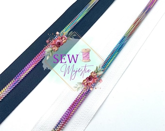 May include: Close-up of two zippers, one navy blue and one white, each with a rainbow-colored metallic zipper tape. The zippers are angled across the frame. The image includes the text "SEW Majestic" with floral accents.