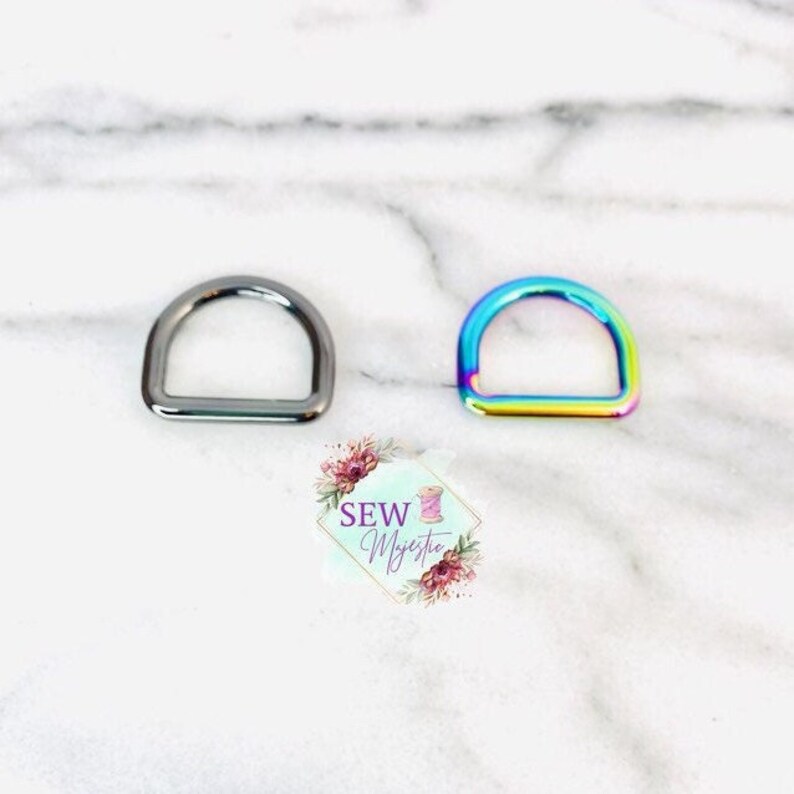 Puede incluir: Dos anillos en D de metal, uno plateado y otro arco&iacute;ris iridiscente. Los anillos est&aacute;n sobre una superficie de m&aacute;rmol blanco.
