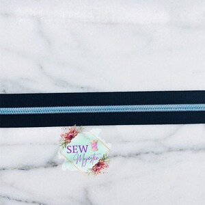 May include: A close-up of a black zipper with a light blue zipper tape. The zipper is on a white marble surface. The text "Sew Majestic" is visible in the lower right corner of the image.
