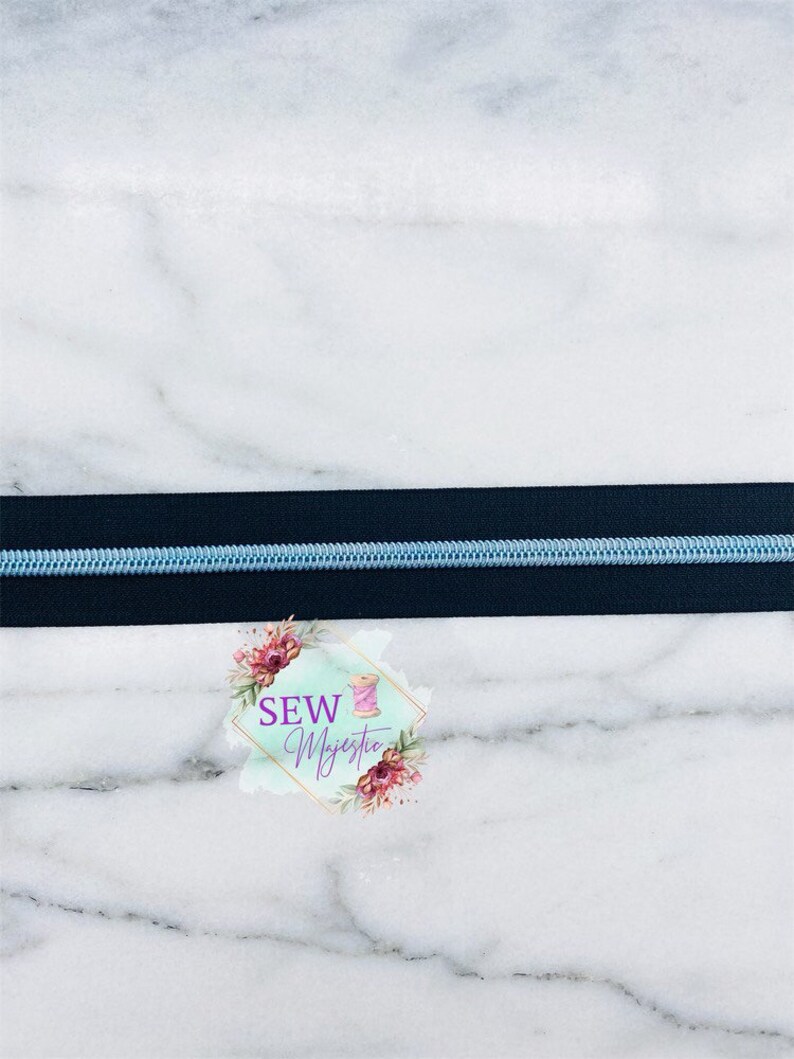 May include: A close-up of a black zipper with a light blue zipper tape. The zipper is on a white marble surface. The text "Sew Majestic" is visible in the lower right corner of the image.
