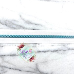May include: A close-up of a white zipper with a light blue zipper tape. The zipper is on a white marble background. The text "SEW Majestic" is visible in the lower center of the image.