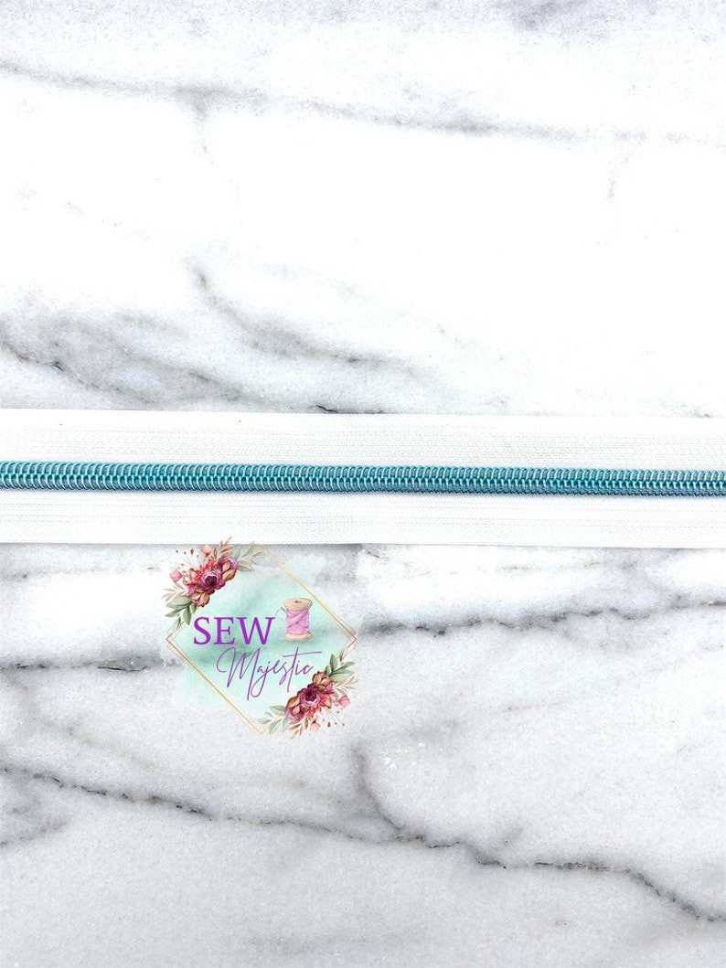 May include: A close-up of a white zipper with a light blue zipper tape. The zipper is on a white marble background. The text "SEW Majestic" is visible in the lower center of the image.