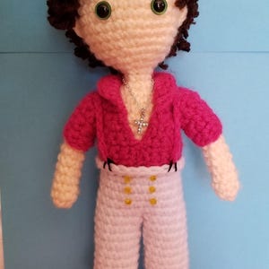 Könnte beinhalten: Gehäkelte Puppe mit braunen Haaren und grünen Augen, trägt ein rosa Hemd und eine weiße Hose mit goldenen Knöpfen. Sie hat eine silberne Kreuzkette und schwarze Schuhe.