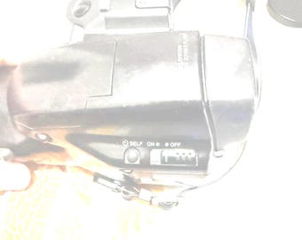 May include: Black digital camera with a zoom lens. The camera has a self-timer, on/off switch, and a flash setting. The camera is held by a person.